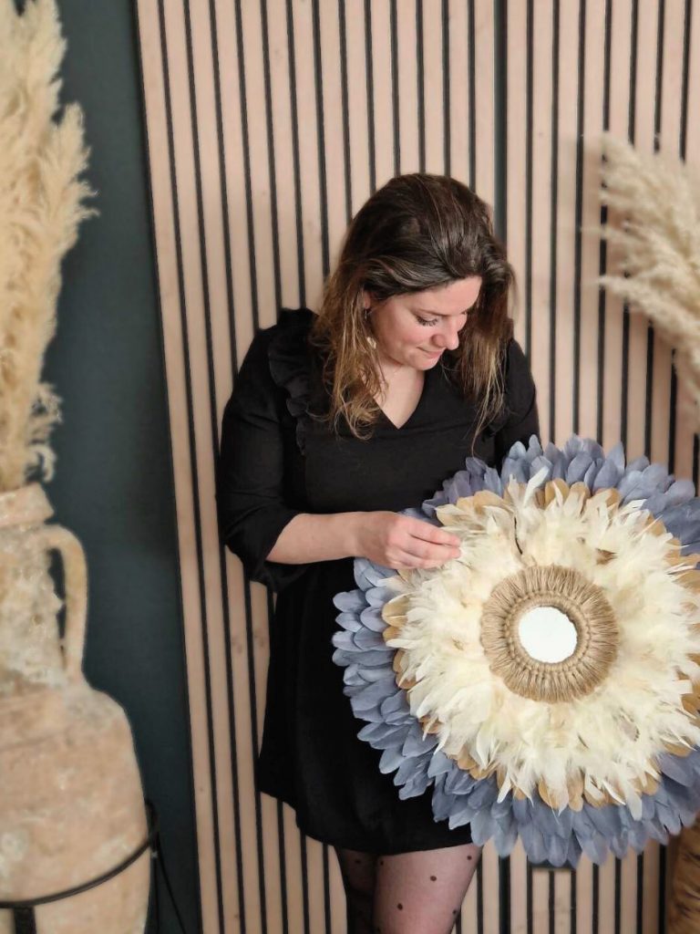 Cliente tenant un miroir en plumes bleu, crème et sable. Décoration bohème artisanale
